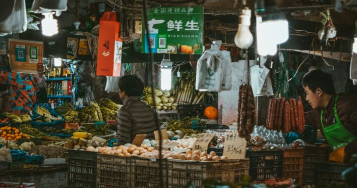 Fotografia em cores de barraca de mercado de rua em Shanghai. É possível ver dois vendedores e um cliente circulando entre caixotes de alimentos.