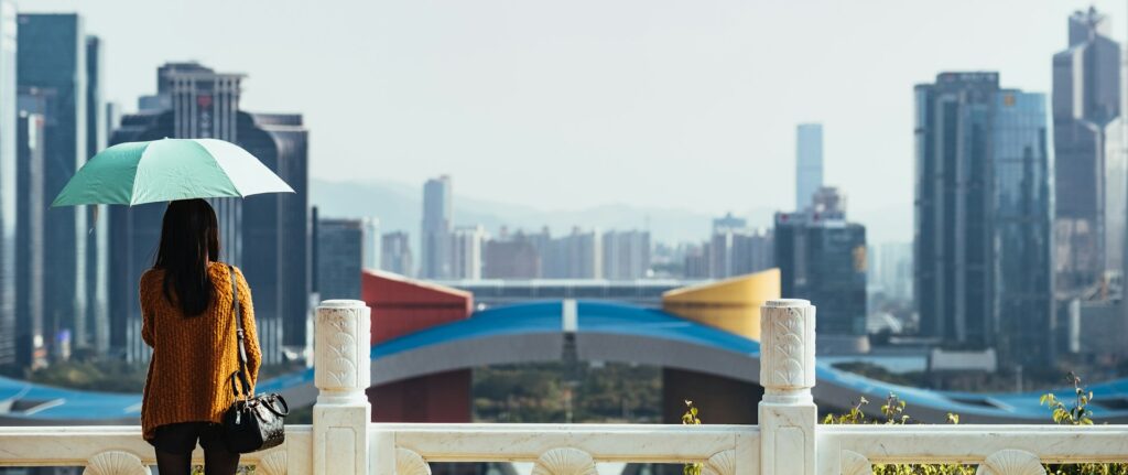 mulher segura um guarda chuva, de costas para a camera, e olha para a paisagem. ela esta em cima de uma ponte com varios predios em frente.