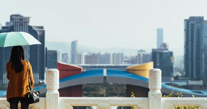 mulher segura um guarda chuva, de costas para a camera, e olha para a paisagem. ela esta em cima de uma ponte com varios predios em frente.