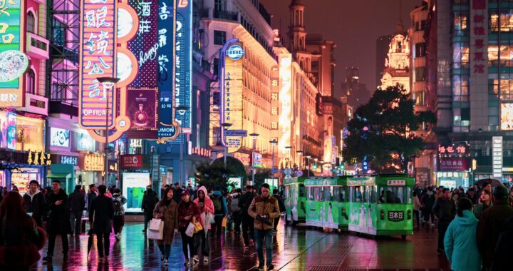 Fotografia em cores de uma rua de Macau à noite com pedestres fazendo compras em meio a luminosos coloridos.