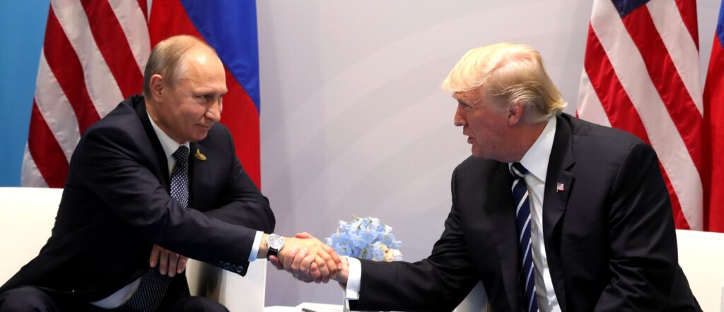 Fotografia em cores de Vladimir Putin e Donald Trump apertando mãos em 2017. Ao fundo, bandeiras dos EUA e da Rússia.