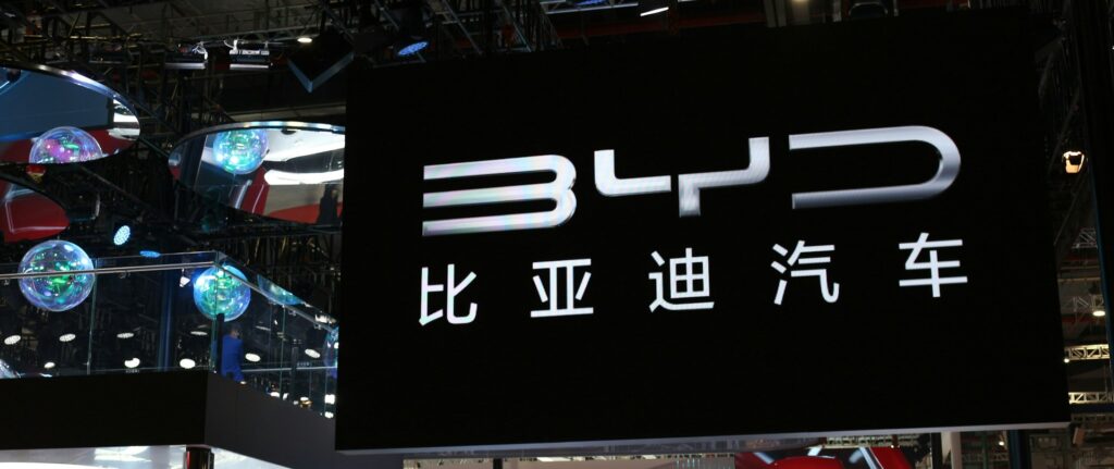 Exposição de carros em um salão com uma placa preta grande escrito BYD e o seu nome em mandarim logo abaixo, em branco, ocupando ⅔ da imagem.