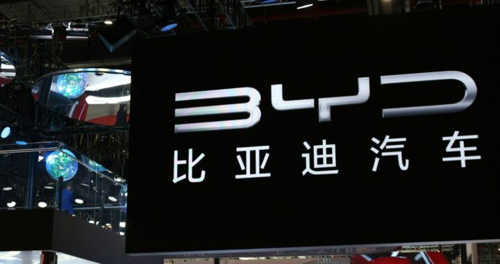 Exposição de carros em um salão com uma placa preta grande escrito BYD e o seu nome em mandarim logo abaixo, em branco, ocupando ⅔ da imagem.