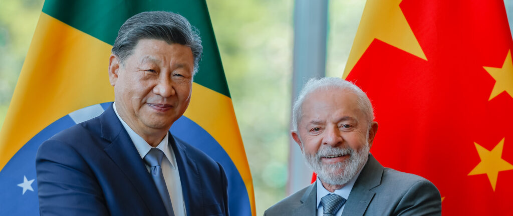 Fotografia em cores de Xi Jinping e Lula sorrindo e apertando mãos. Ao fundo, uma bandeira do Brasil (atrás de Xi) e uma bandeira da China (atrás de Lula). A foto é de 2024.