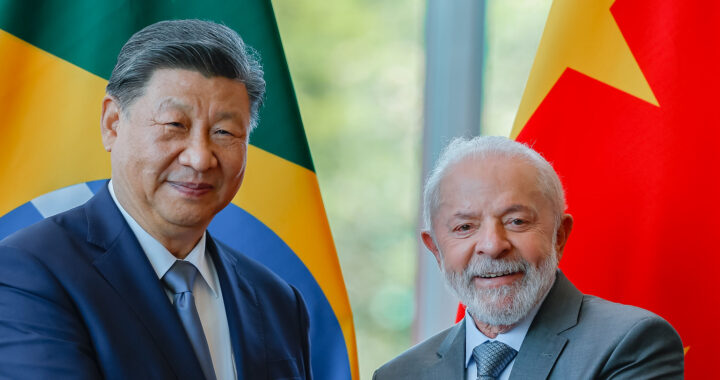 Fotografia em cores de Xi Jinping e Lula sorrindo e apertando mãos. Ao fundo, uma bandeira do Brasil (atrás de Xi) e uma bandeira da China (atrás de Lula). A foto é de 2024.