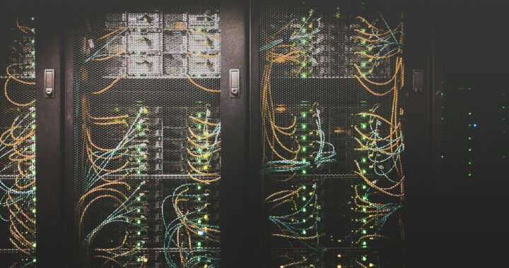 Fotografia em cores de um armário em um data center. Dentro dele, é possível ver cabos amarelos e verdes, além de leds acesos.