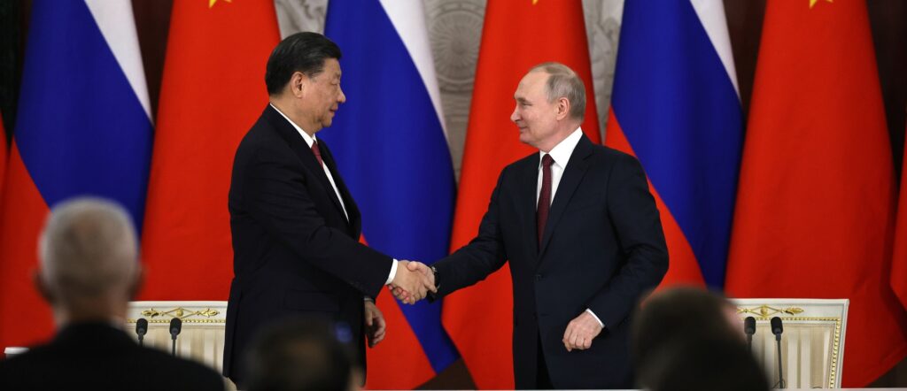 Fotografia em cores de Xi Jinping e Vladimir Putin em aperto de mãos em 2023. Ao fundo, bandeiras da China e da Rússia.