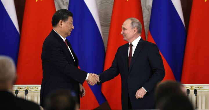 Fotografia em cores de Xi Jinping e Vladimir Putin em aperto de mãos em 2023. Ao fundo, bandeiras da China e da Rússia.
