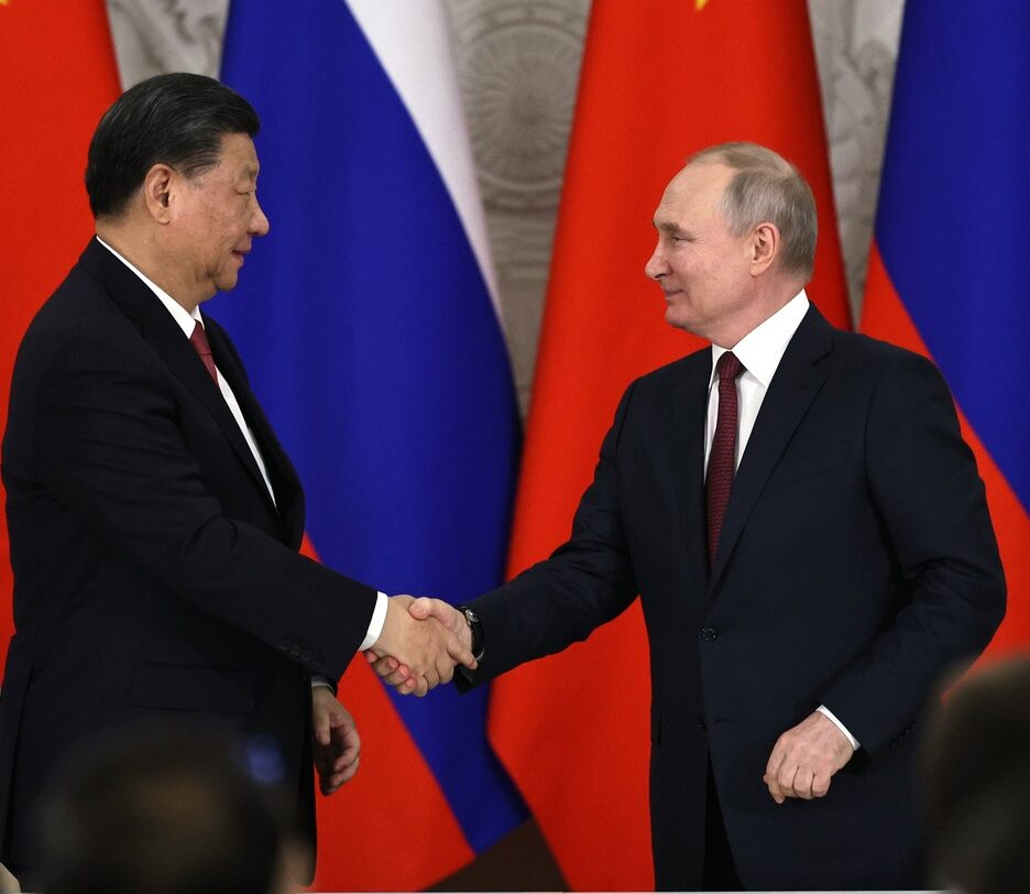 Fotografia em cores de Xi Jinping e Vladimir Putin em aperto de mãos em 2023. Ao fundo, bandeiras da China e da Rússia.