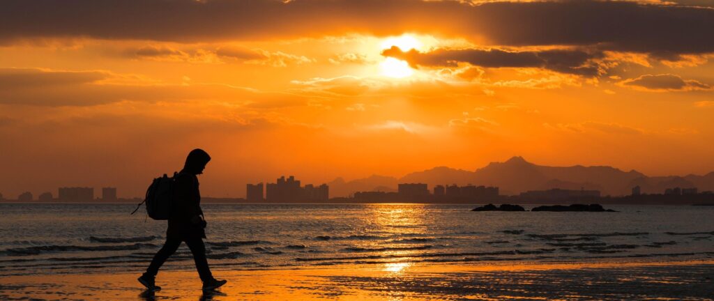 Fotografia em cores de pôr do sol em praia em Beidaihe. Em primeiro plano, há o perfil de uma pessoa caminhando. Ao fundo, é possível ver algumas construções.