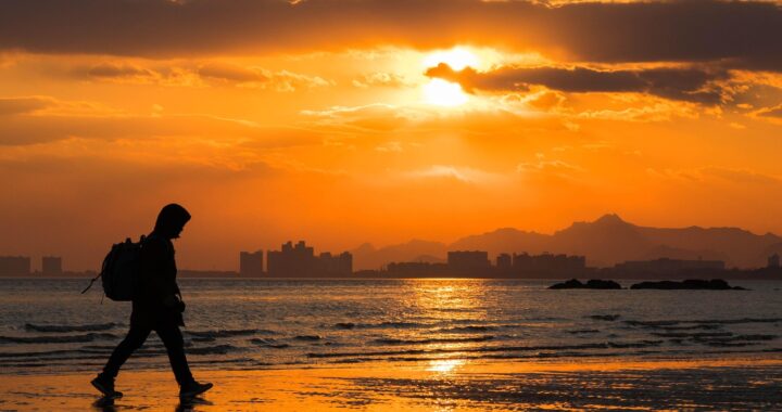 Fotografia em cores de pôr do sol em praia em Beidaihe. Em primeiro plano, há o perfil de uma pessoa caminhando. Ao fundo, é possível ver algumas construções.