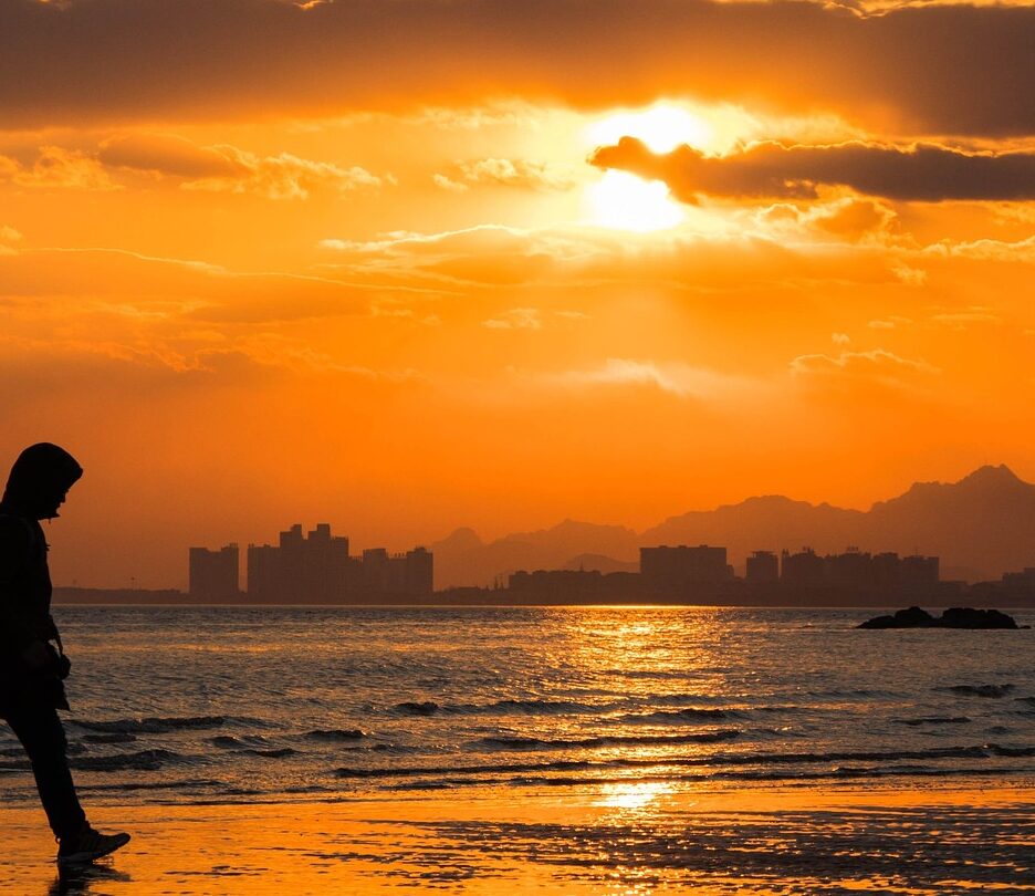 Fotografia em cores de pôr do sol em praia em Beidaihe. Em primeiro plano, há o perfil de uma pessoa caminhando. Ao fundo, é possível ver algumas construções.