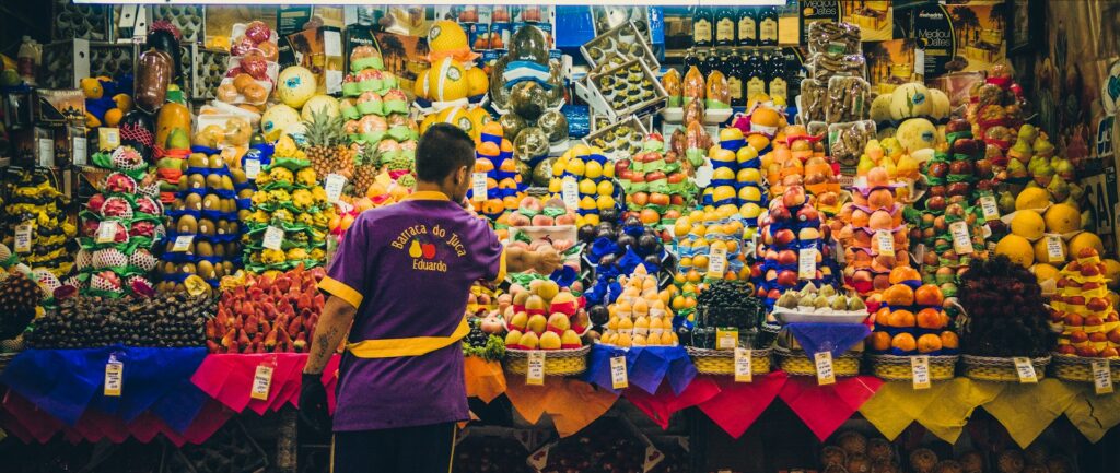 Fotografia em cores de vendedor, de costas, diante de uma barraca de feira cheia de frutas.