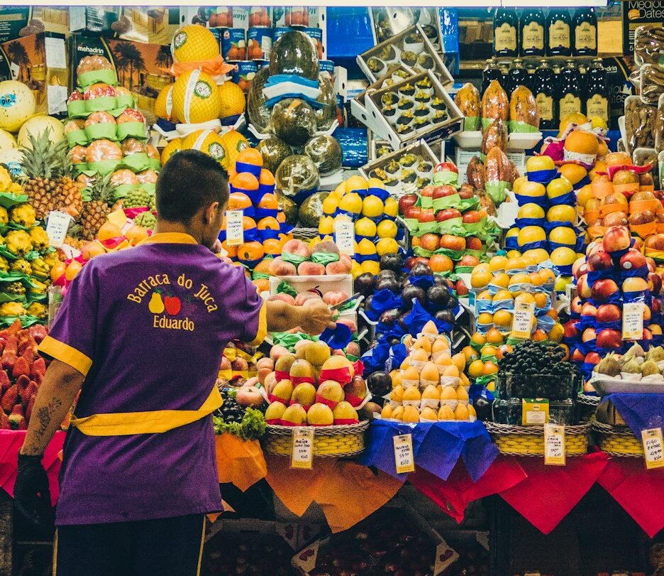 Fotografia em cores de vendedor, de costas, diante de uma barraca de feira cheia de frutas.