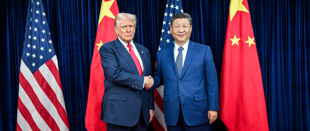 Fotografia em cores de Donald Trump e Xi Jinping em outubro de 2025. Eles posam diante das bandeiras de seus países, apertando as mãos. Xi sorri, mas Trump faz cara feia.