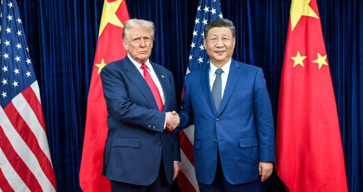 Fotografia em cores de Donald Trump e Xi Jinping em outubro de 2025. Eles posam diante das bandeiras de seus países, apertando as mãos. Xi sorri, mas Trump faz cara feia.