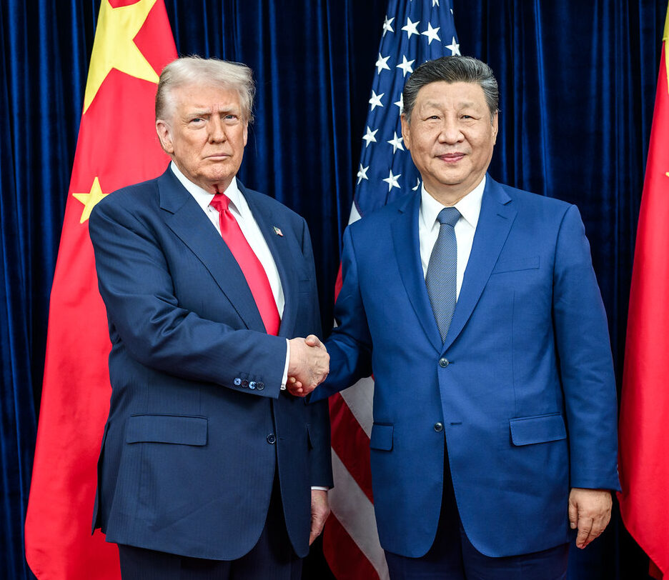 Fotografia em cores de Donald Trump e Xi Jinping em outubro de 2025. Eles posam diante das bandeiras de seus países, apertando as mãos. Xi sorri, mas Trump faz cara feia.