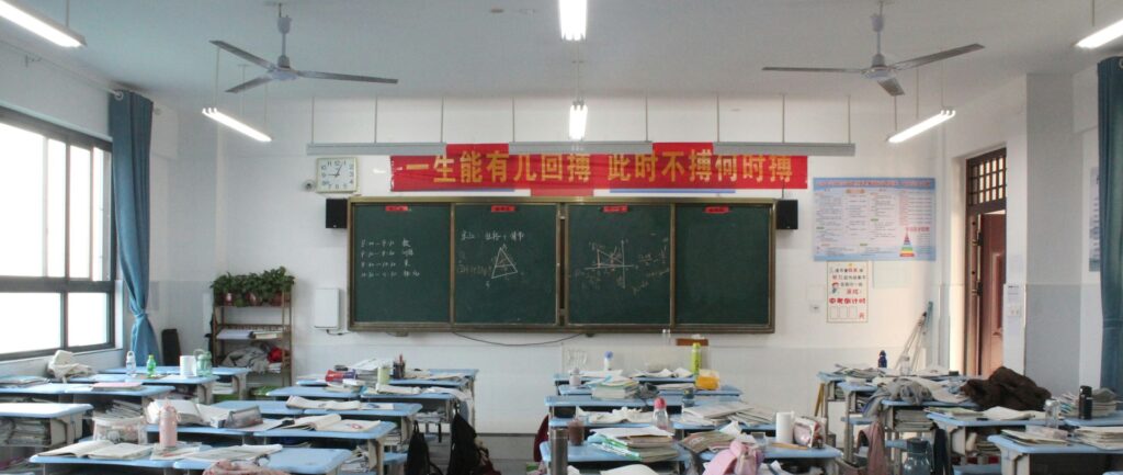 Fotografia em cores de uma sala de aula chinesa sem muita tecnologia. São visíveis a lousa de giz, carteiras, cadernos e outros objetos pessoais de alunos, que não aprecem na foto.