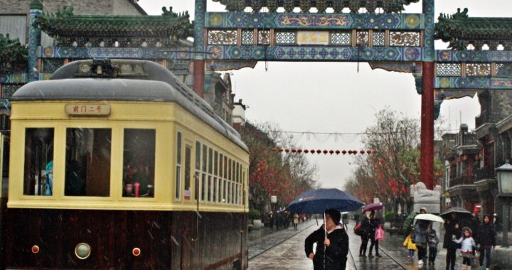 Fotografia em cores de bonde de Pequim, amarelo e marrom, circulando sob um pórtico em estilo chinês. Chove, é possível ver algumas pessoas usando guarda-chuvas.
