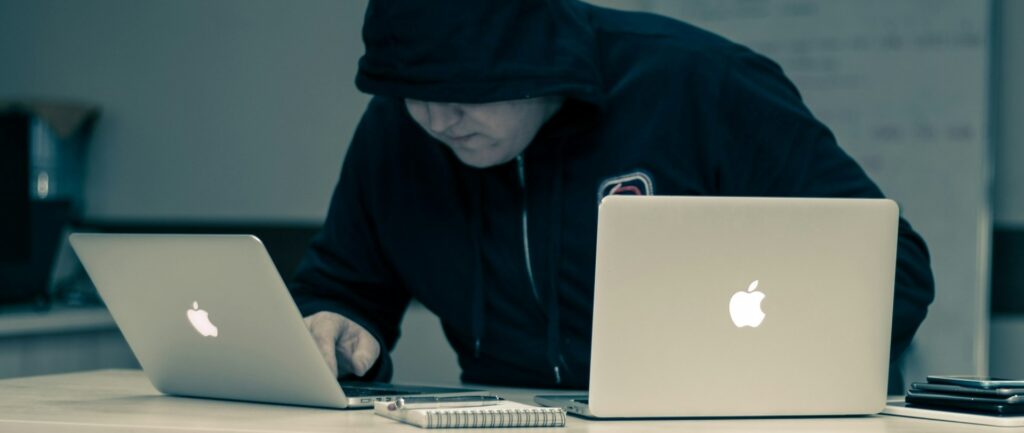 foto em preto em branco mostra homem com rosto semi coberto por capuz preto enquanto manipula dois computadores com a logo da Apple em destaque