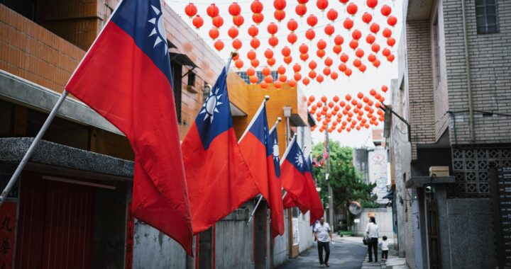 Foto em cores de sequência de bandeiras de Taiwan em uma rua com pedestres e com lanternas chinesas ao fundo.