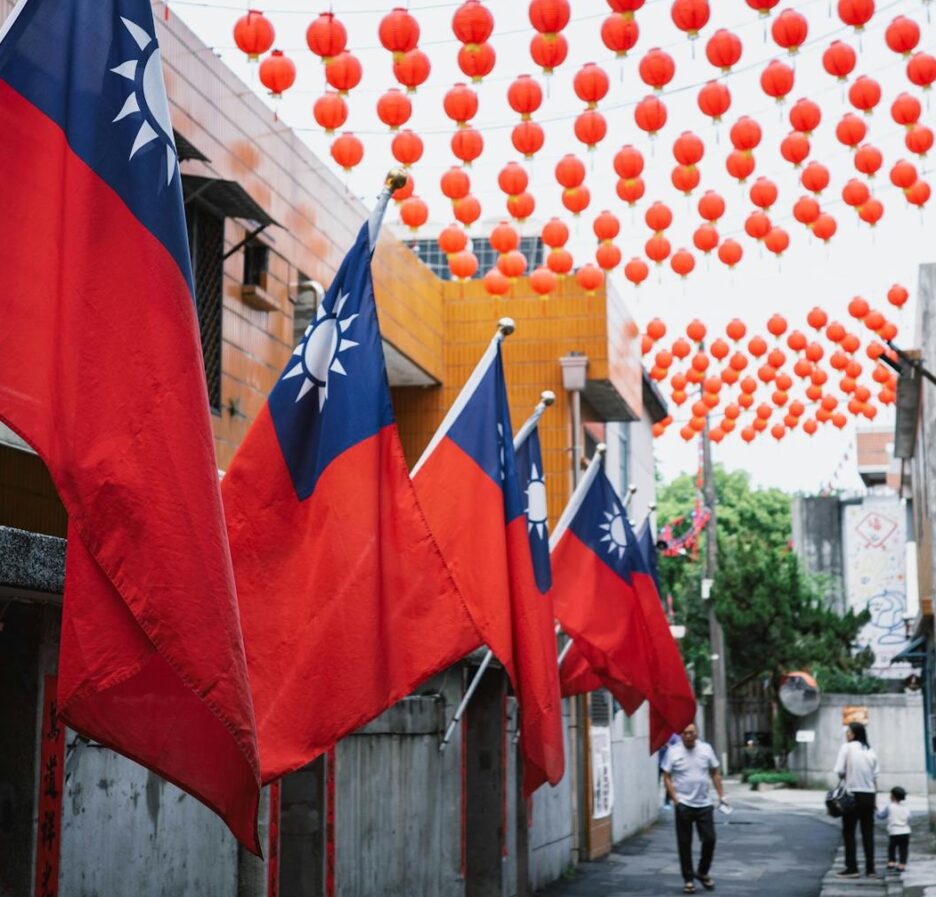 Foto em cores de sequência de bandeiras de Taiwan em uma rua com pedestres e com lanternas chinesas ao fundo.