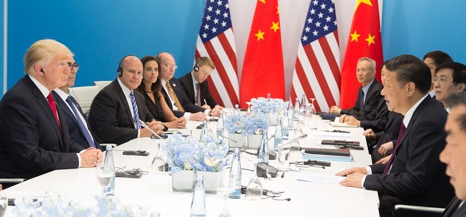 mesa de reunião com Trump e equipe sentados de um lado e do outro Xi Jinping e equipe, com bandeiras no fundo