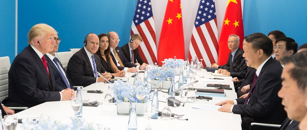 mesa de reunião com Trump e equipe sentados de um lado e do outro Xi Jinping e equipe, com bandeiras no fundo