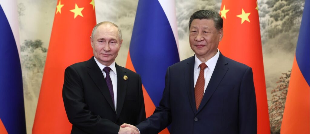 Fotografia em cores de Vladimir Putin e Xi Jinping apertando as mãos. Bandeiras da China e da Rússia estão ao fundo.