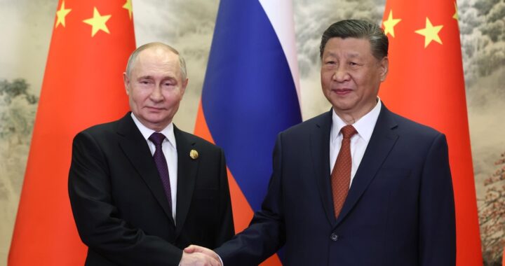 Fotografia em cores de Vladimir Putin e Xi Jinping apertando as mãos. Bandeiras da China e da Rússia estão ao fundo.