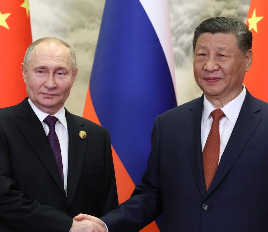 Fotografia em cores de Vladimir Putin e Xi Jinping apertando as mãos. Bandeiras da China e da Rússia estão ao fundo.