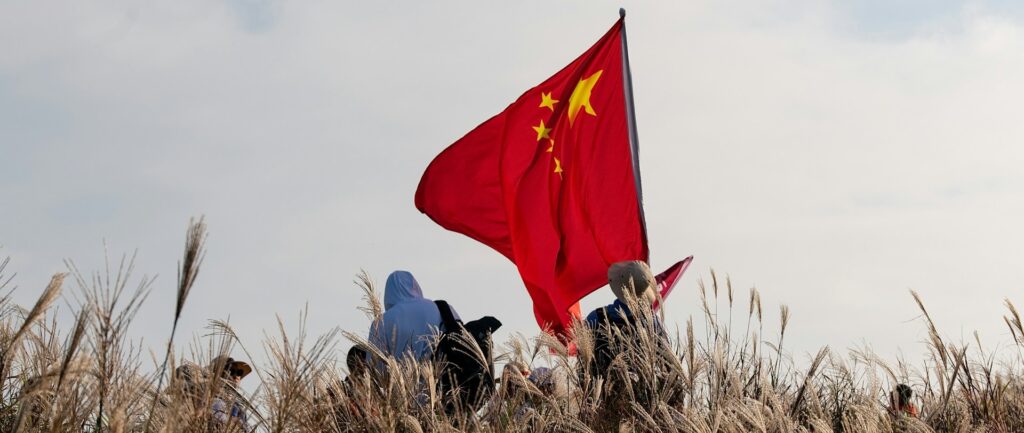 Fotografia em cores de pessoas, encobertas por grama alta, segurando uma grande bandeira da China.