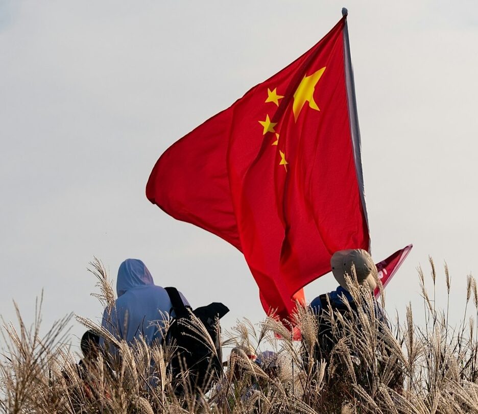 Fotografia em cores de pessoas, encobertas por grama alta, segurando uma grande bandeira da China.
